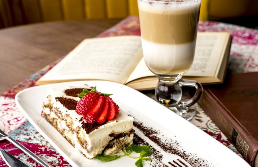 side view of tiramisu decorated with sliced strawberries and cocoa powder on a plate served with a glass of latte macchiato on the table
